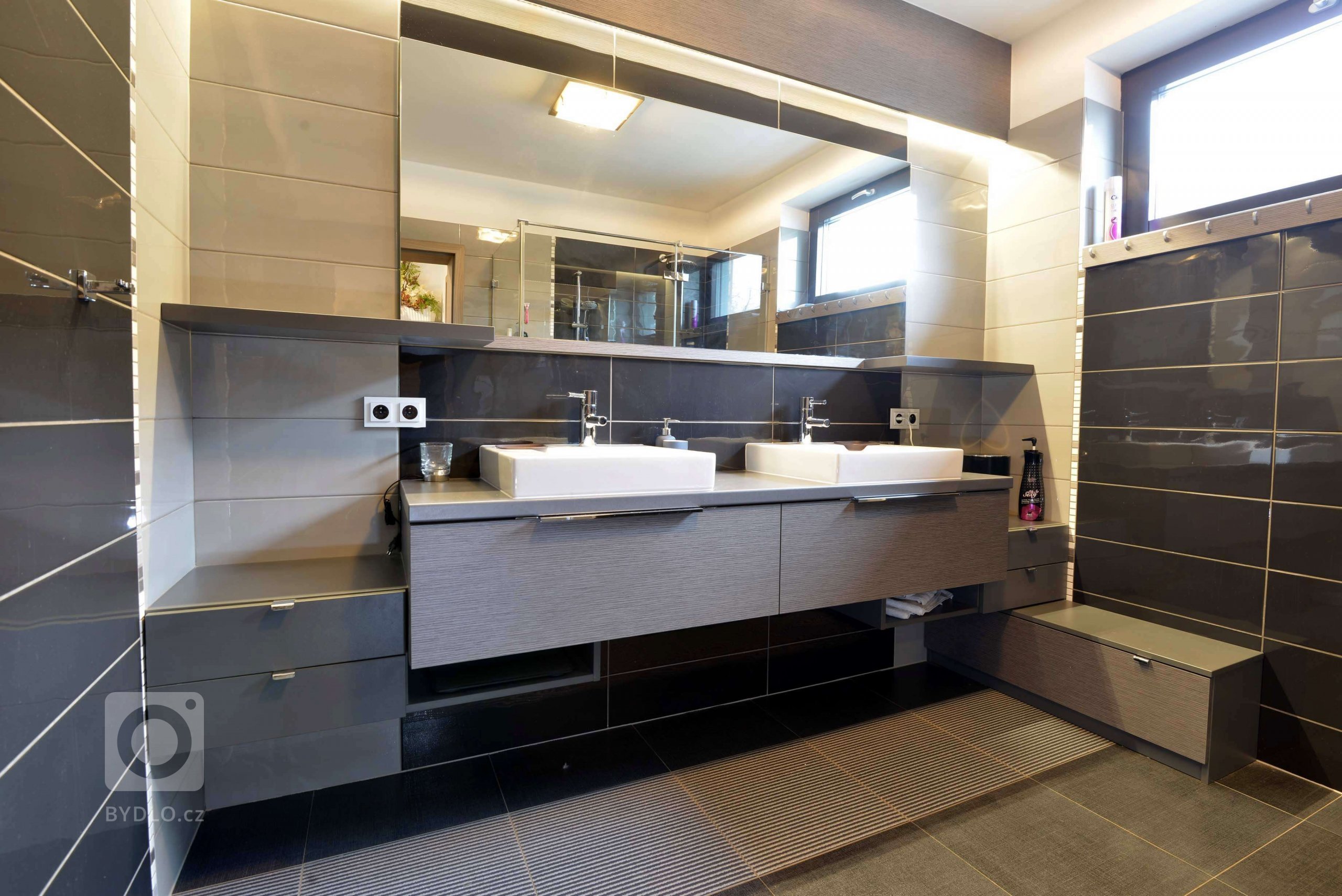 Koupelna pro rodiče. Vše je v odstínech šedé a černé. Toto je pohled na koupelnový nábytek s dvěma umyvadly, každý z páru má svojí stranu. Sestavu doplňuje velkoplošné zrcadlo a lišta s osvětlením LED. 