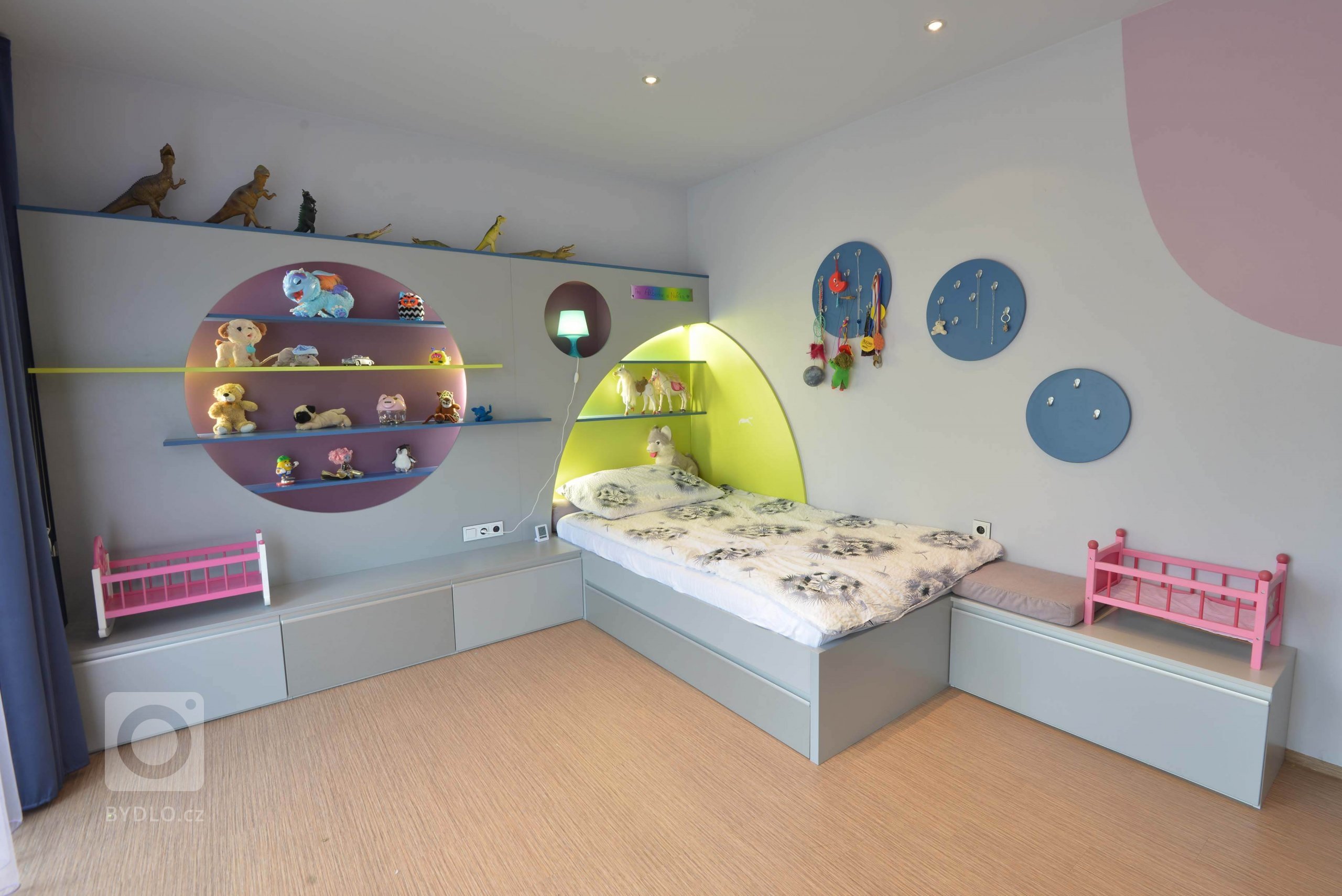 Pohled na dělanou postel s úložným prostorem. Pokoji vévodí předstěna s kulatými a půlkulatými výřezy na odkládání hraček. Vše se zabudovaným osvětlením LED.