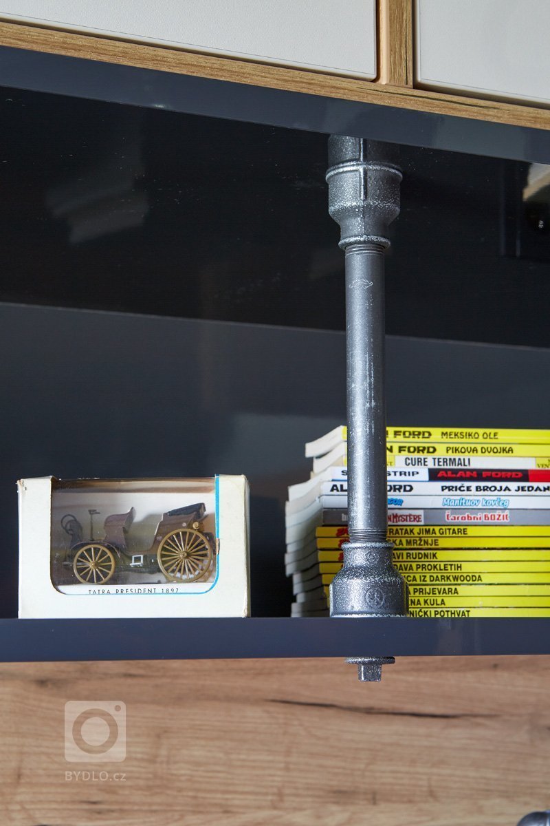 Knihy našly místo na dlouhé polici, kde je i zásuvka na dobíjení mobilních zařízení. Případnému prohnutí brání dvě trubky, které jsou zároveň i designovým prvkem a spolu s lampičkami, výrobními na zakázku dle autorského návrhu, dávají celé místnosti industriální nádech. 