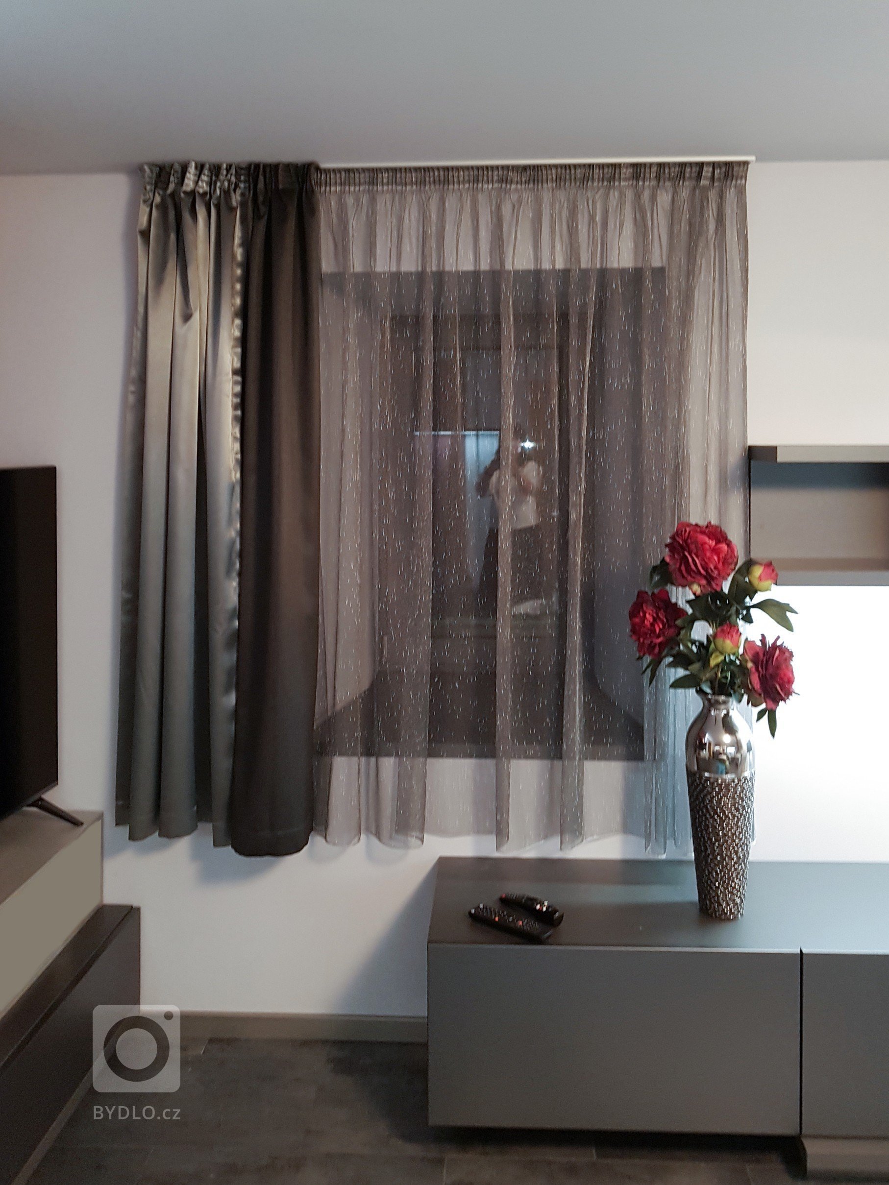 Detai závěsů, které jsou kombinované ze dvou materiálů ve stříbrné barvě v matu a lesku a záclona s nitkami ve stříbrné metalické. 