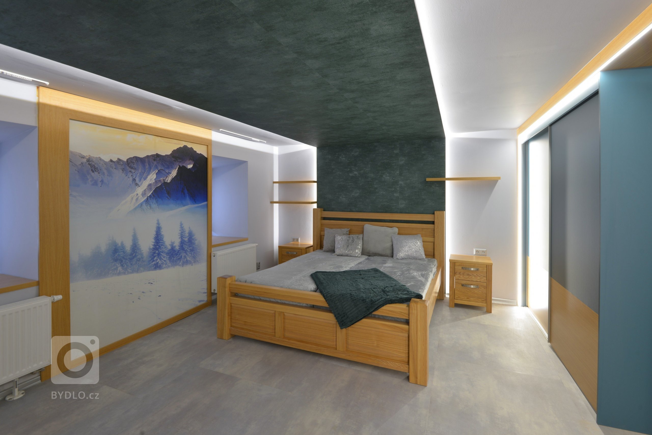 Středem prostoru vede snížený sádrokartonový strop, který na obou stranách přechází do předstěny, na jedné straně tvoří pozadí k posteli se skrytým led osvětlením, na protilehlé straně je jako pozadí pro sedací kout a prostor na zavěšenou televizi, která tam v době focení ještě nebyla. Uprostřed místnosti mezi okny je obrazová tapeta s horskými motivy.