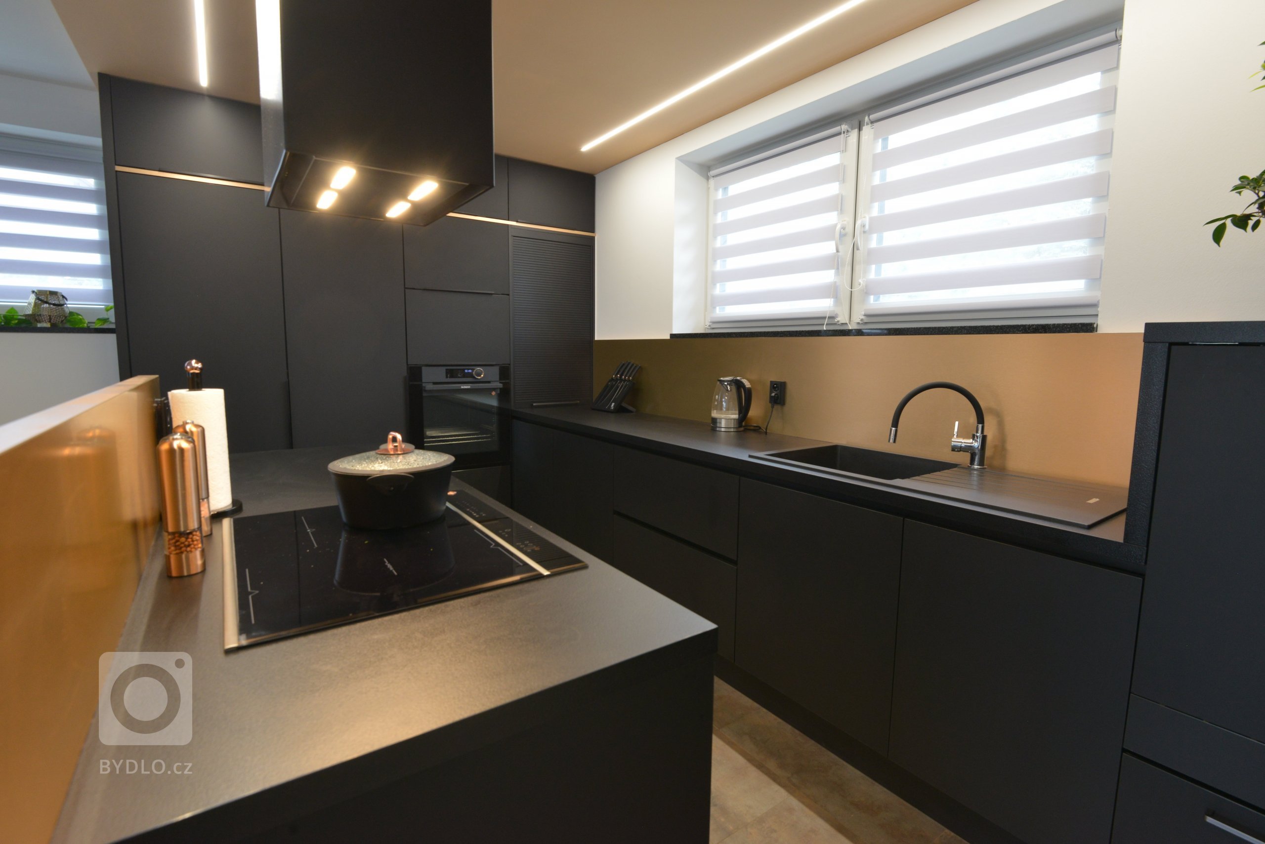 Pracovní plocha pod oknem umožňuje výhled a přitom má koncentrované mycí centrum a zvýšenou myčku. V rohu je černá roletová skříňka na domácí spotřebiče.