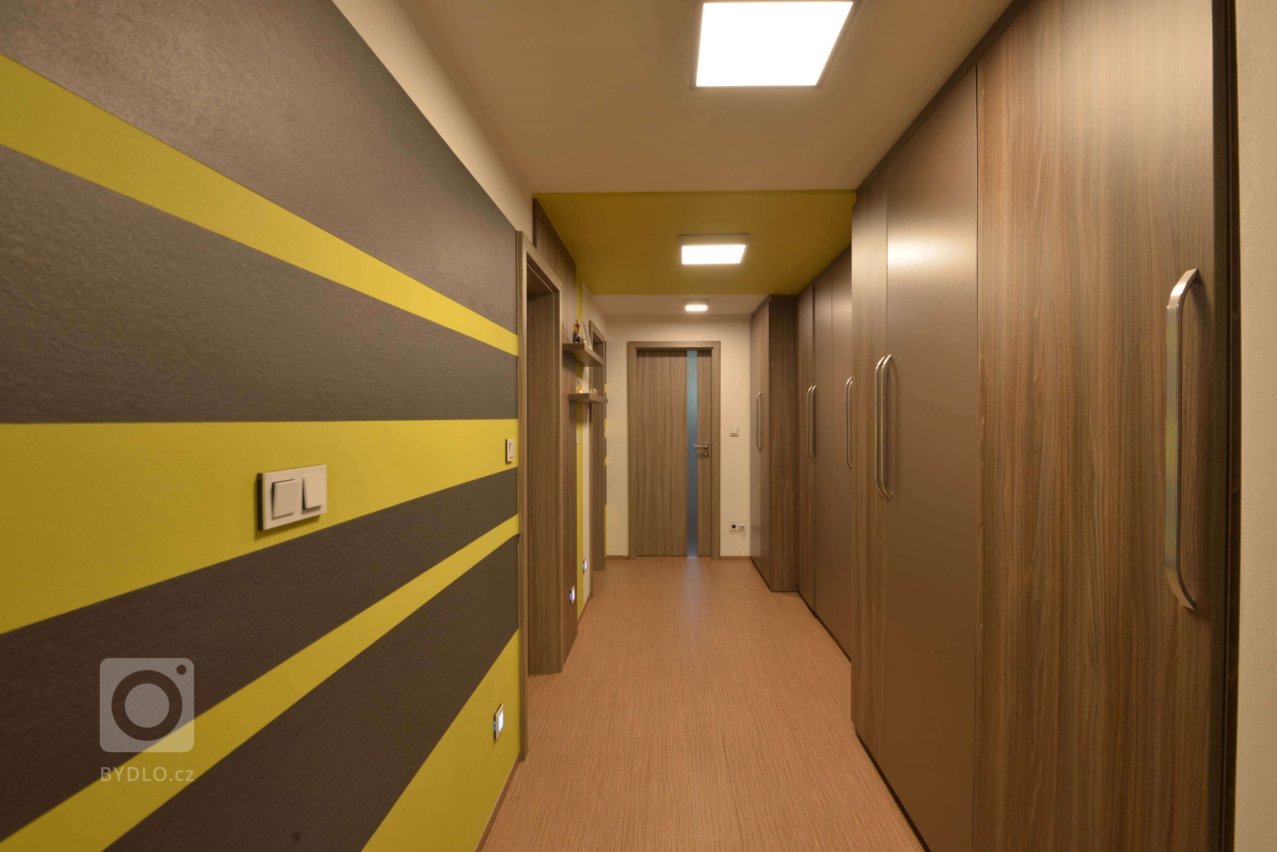 Chodba je doladěna pruhy z antracitové tapety a žlutozelené výmalbě. Část výmalby prochází přes strop. V chodbě u dětských pokojů je výmalba podložená magnetickým nátěrem, takže vytváří variabilní nástěnku pro děti, na vystavení čehokoliv. 