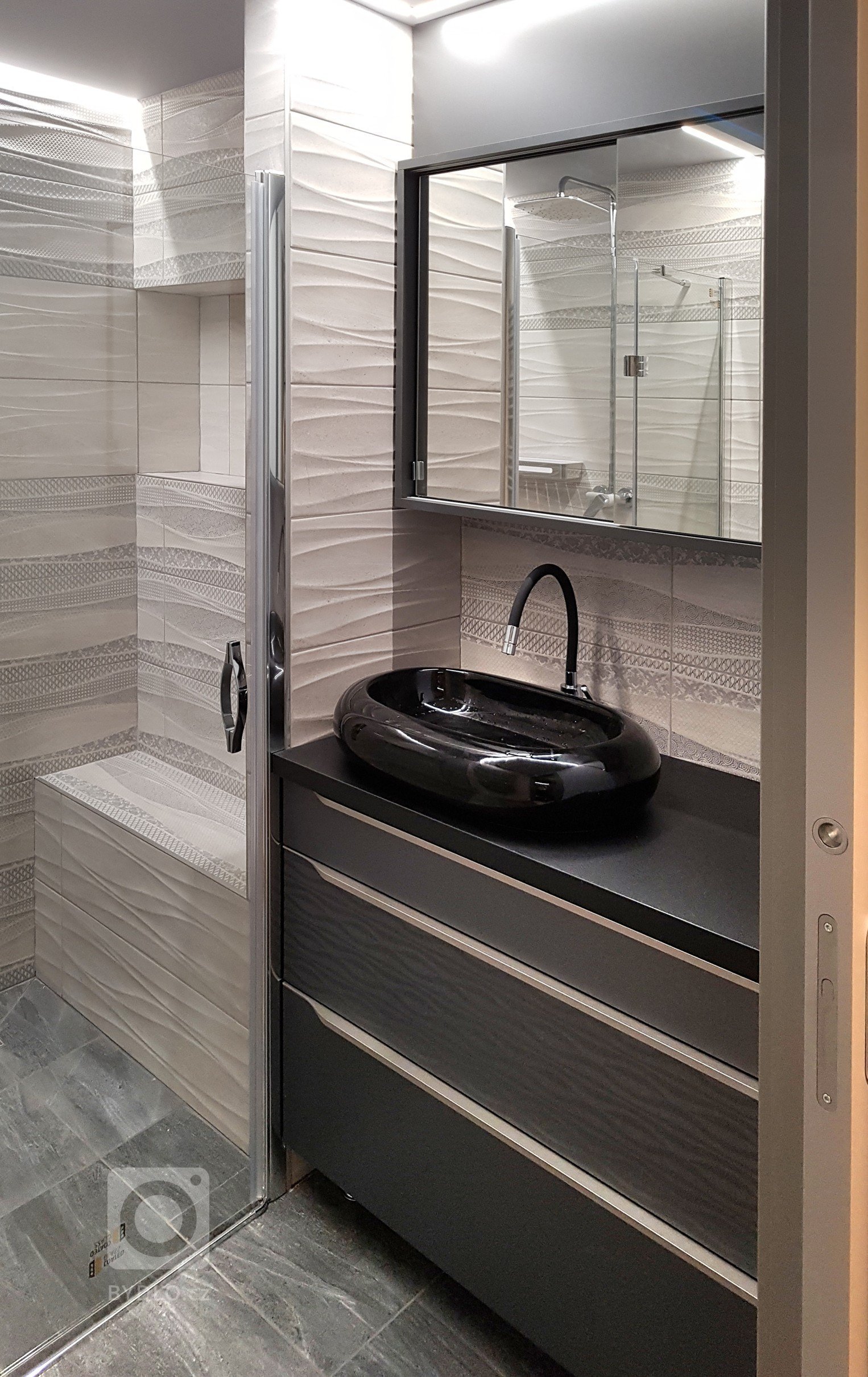 Malá koupelna z prostoru na konci úzké chodby. V koupelně je přes celou šíři sprchový kout s vyzděnou lavičkou a odkládacím prostorem na šampony aj.