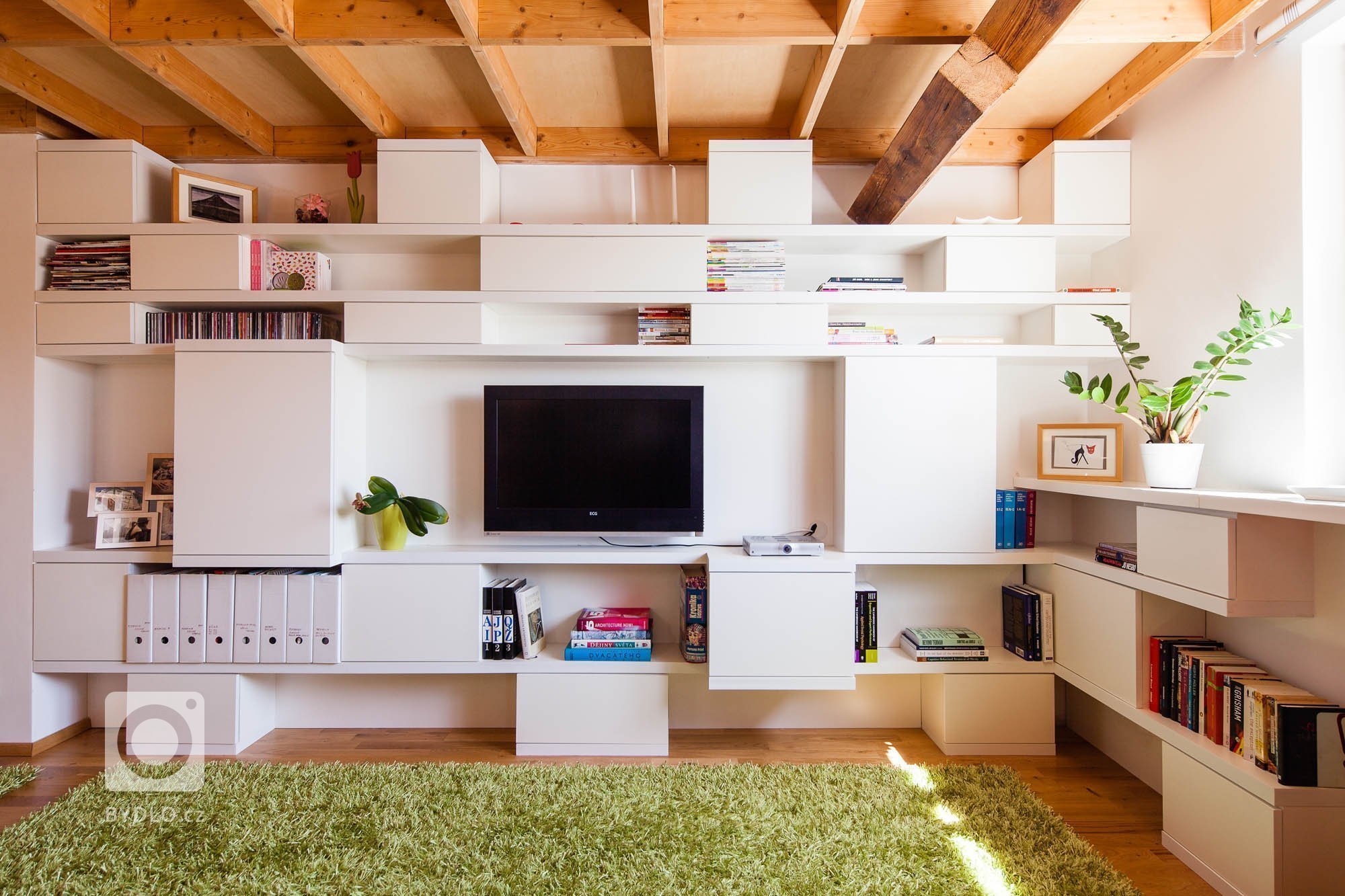 Knihovna je vložena do trámoví podkrovního bytu, odděluje prostor obývacího pokoje a kuchyně, ale zároveň ponechává vizuální kontakt. Ortogonální rastr otvorů…