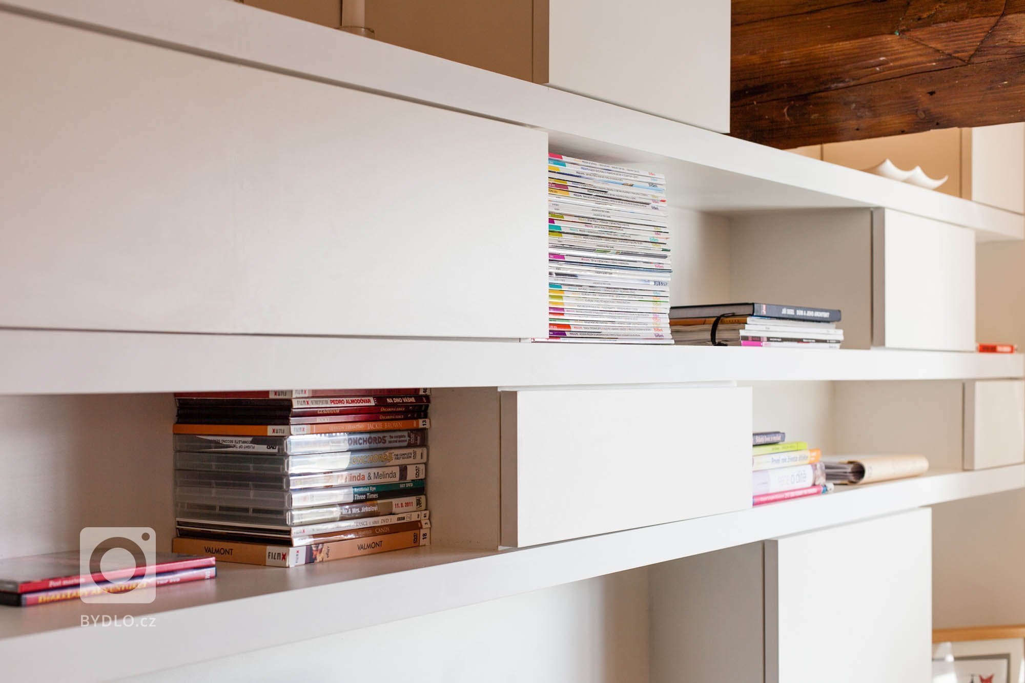 Knihovna je vložena do trámoví podkrovního bytu, odděluje prostor obývacího pokoje a kuchyně, ale zároveň ponechává vizuální kontakt. Ortogonální rastr otvorů…