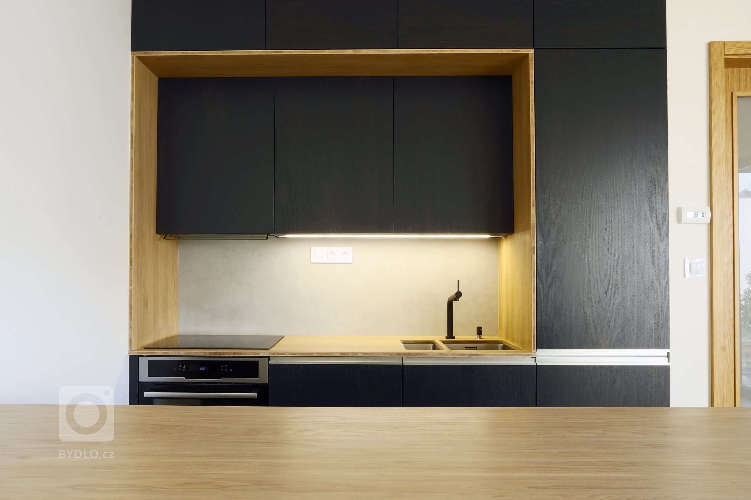 Odvážná kuchyně v novostavbě, kdy je tmavá barva zjemněná dubovou bio deskou a kresba dřeva pod dýhovaným mořeným dvířkem krásně prostupuje a vytváří zajímavý…