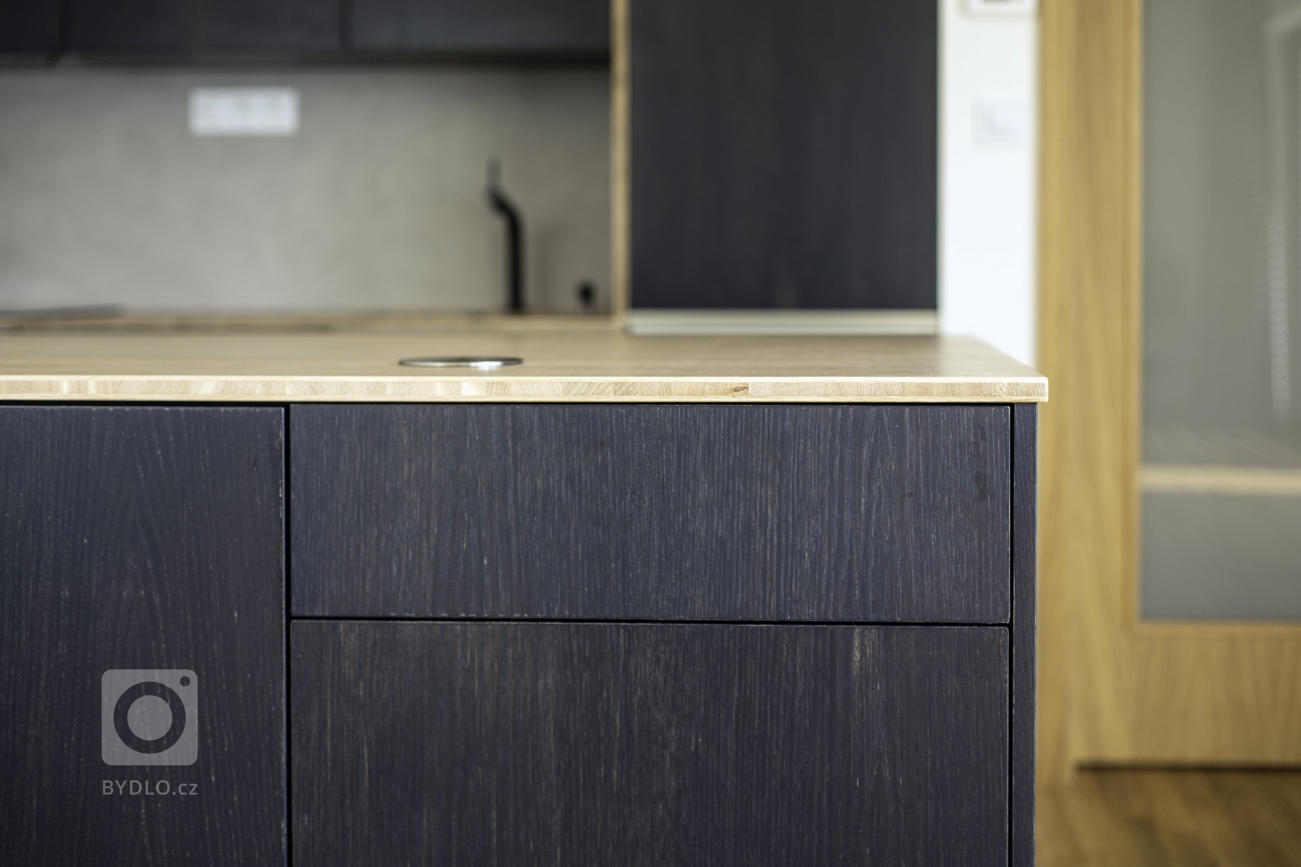 Odvážná kuchyně v novostavbě, kdy je tmavá barva zjemněná dubovou bio deskou a kresba dřeva pod dýhovaným mořeným dvířkem krásně prostupuje a vytváří zajímavý…