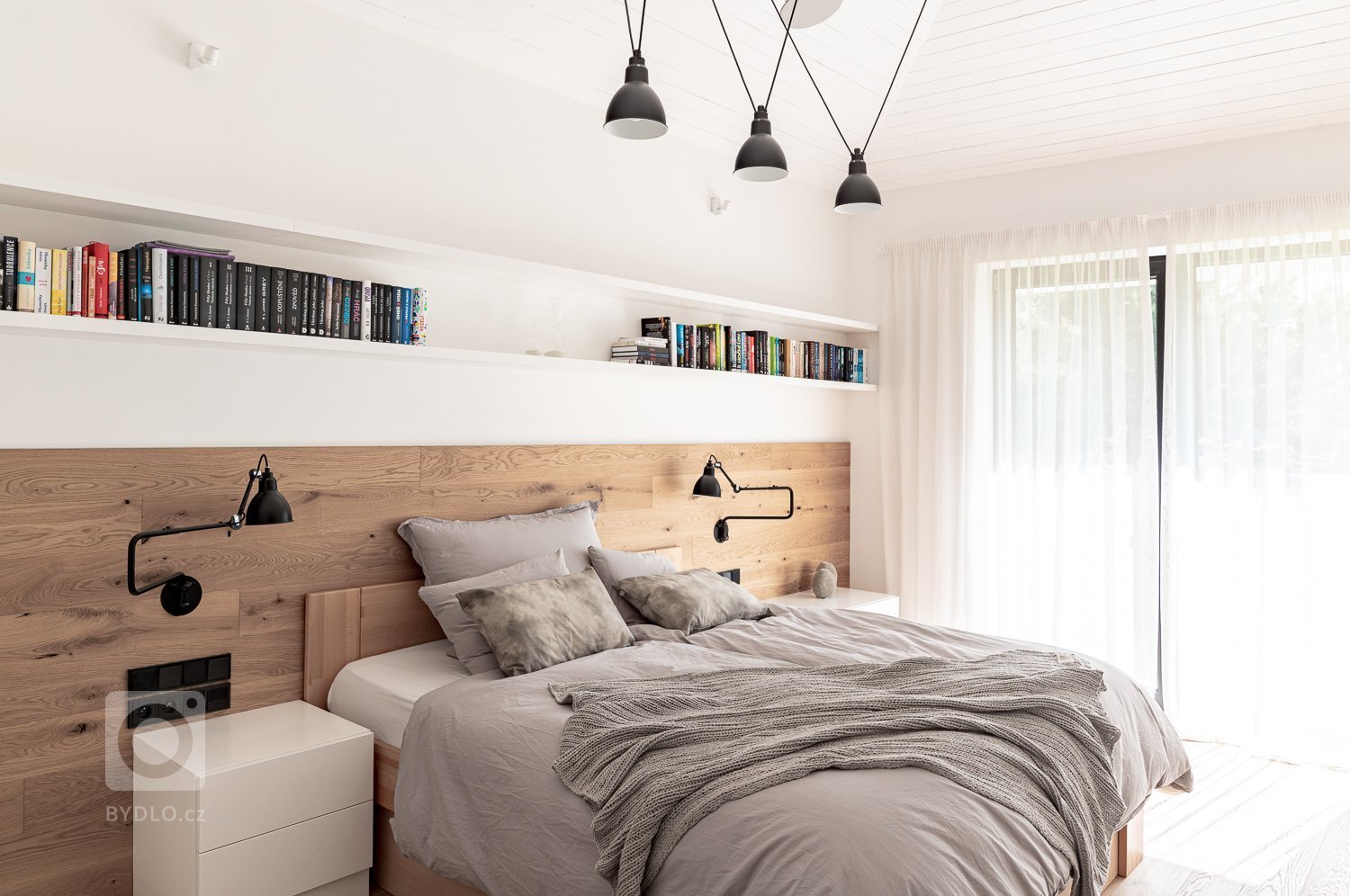 Dřevěná podlaha přechází v obložení stěny, která tak podtrhuje samotnou postel vůči protilehlé nábytkové stěně.