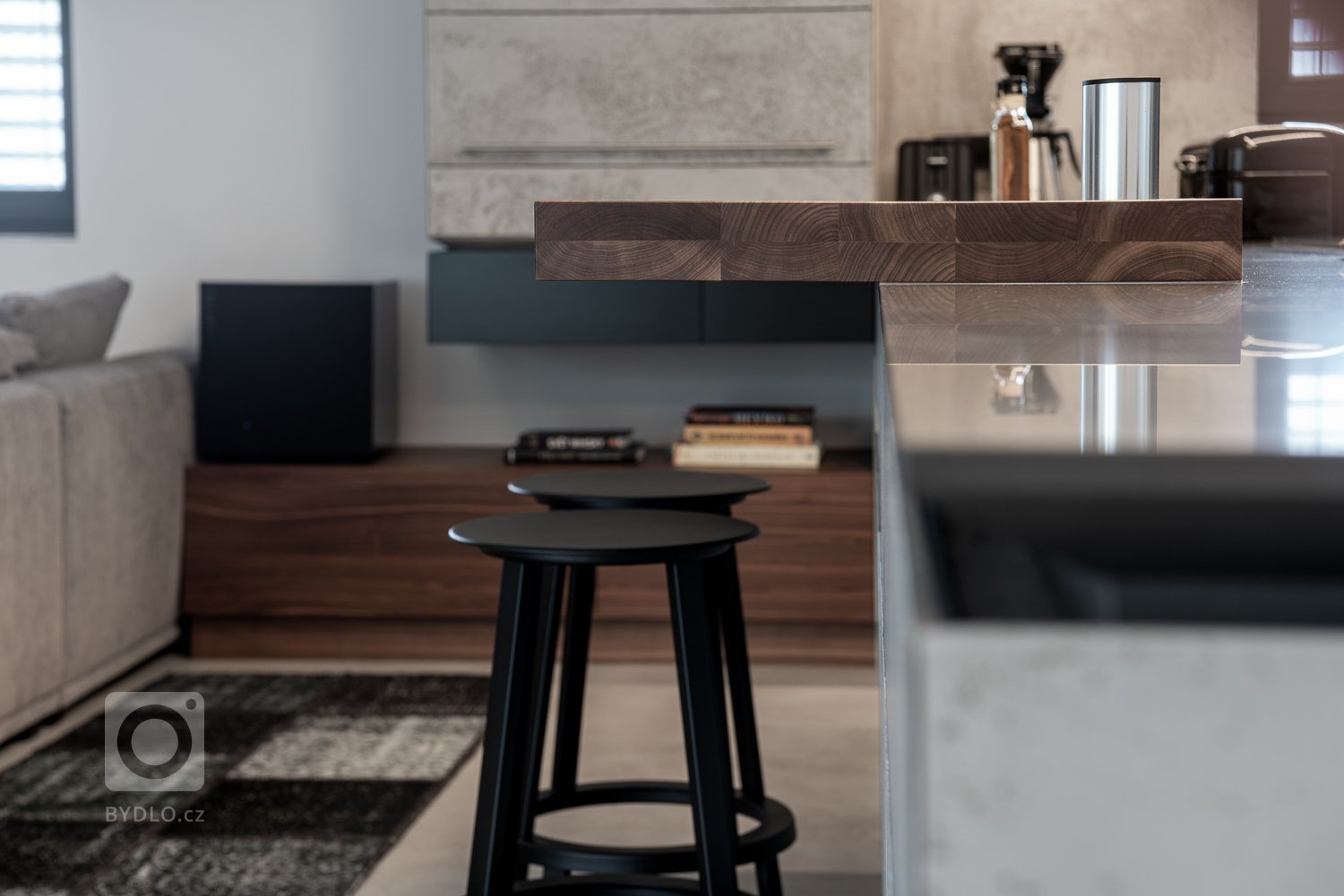 Subtilní pracovní deska z umělého kamene se dostává do kontrastu s masivní barovou deskou z ořechu, s místy k sezení pro dvě osoby. 