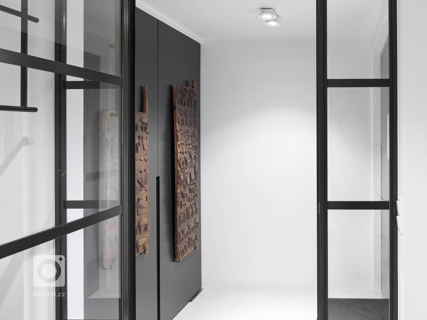 Velké pivotové dveře na celou výšku místnosti dominují vstupu a poskytují rám (jako obraz) celé místností. 