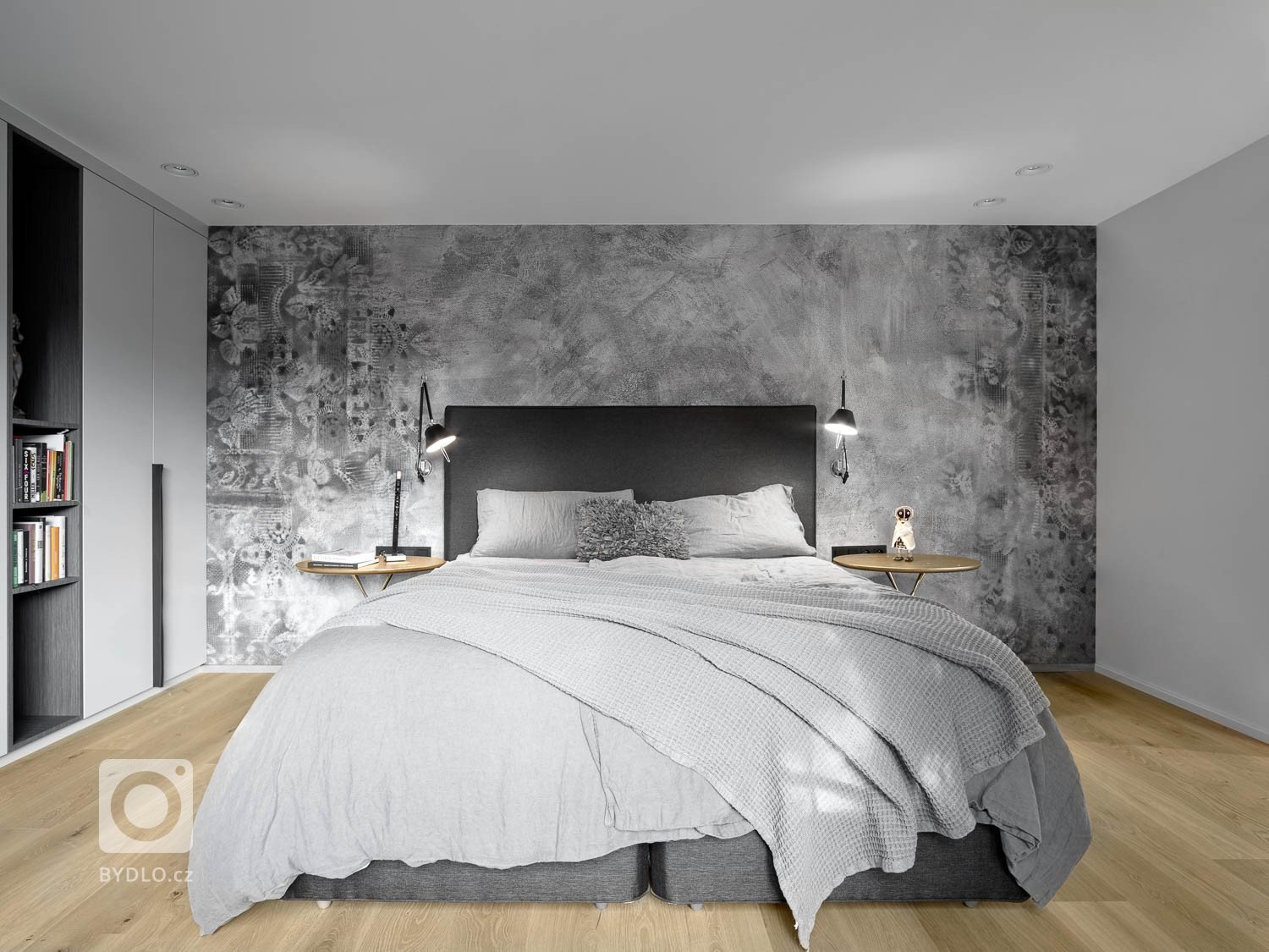 Ložnice má vestavné šatní skříně z lakované MDF, ve světle šedé barvě, doplněné o kožené úchytky. Stěna je dekorována tapetou se vzorem od Wall and Deco, na níž vyniká antracitová čalouněná kontinentální postel. 