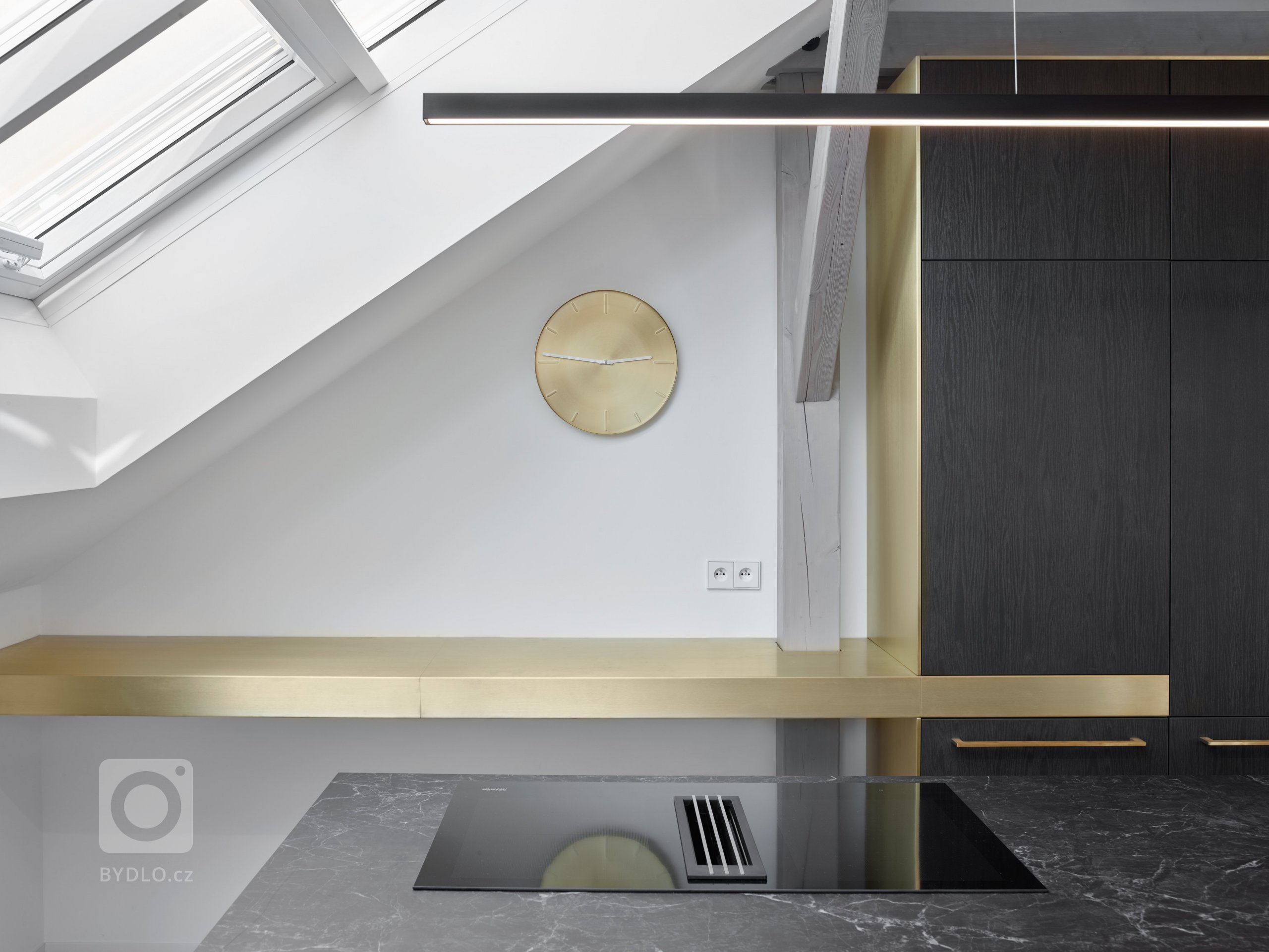 Zapracováním kuchyně do otevřeného krovu vznikla geometricky vyvážená kompozice, která je nejen estetická, ale i praktická díky přirozenému světlu dopadající z podkrovních oken. Kuchyňská linka z tmavé dýhy kontrastuje s mosaznými prvky jako je kování a odkládací deska.