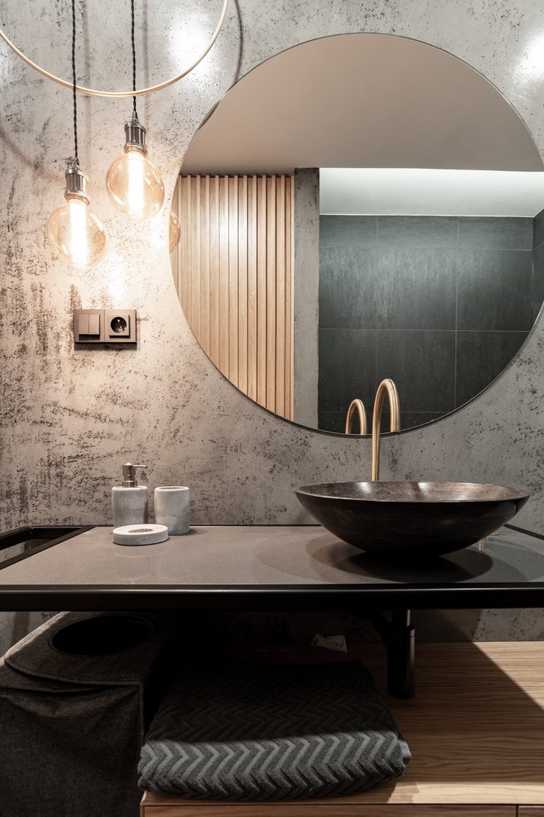 V koupelně u ložnice se jedná o zrcadlo podsvícené, v hlavní koupelně je doplněno o designový autorský prvek mosazné obruče, kombinované se závěsnými Edison žárovkami. 