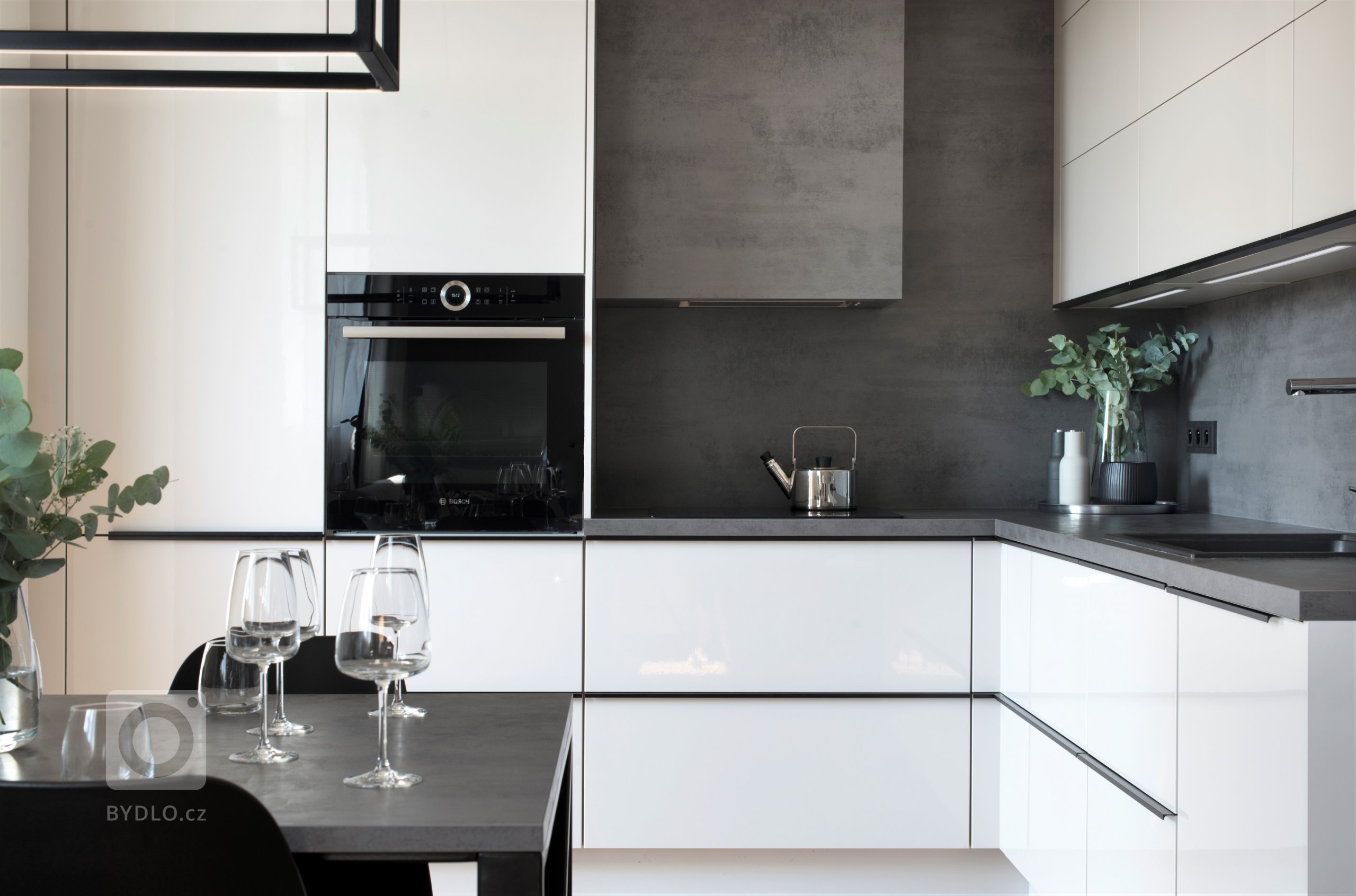 Kuchyně v kombinaci světlých lesklých dvířek s tmavě šedou pracovní deskou, doplněná kontrastní černou úchytkou.
.
Celý prostor propojuje dekor pracovní desky, který se prolíná i na jídelním a konferenčním stolu.
.
Narážecí úchytky, které procházejí po horní hraně dvířek, nenarušují celkový vzhled kuchyně a podtrhují její čisté linie.