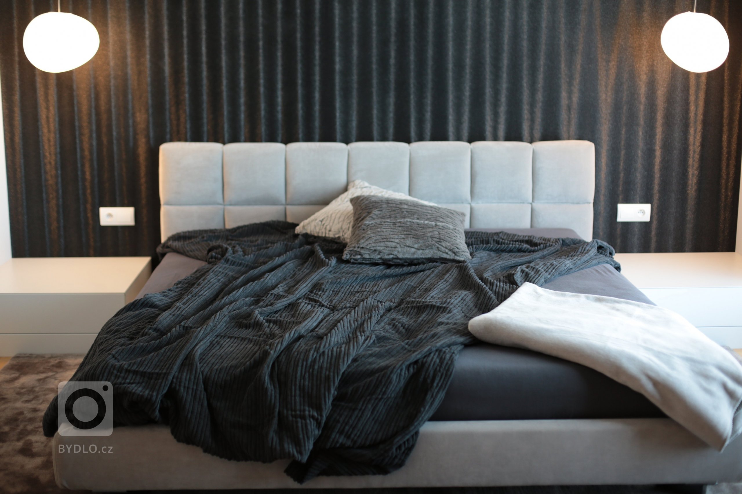 Celočalouněná postel je umístěna na malém pódiu v nice, jehož součástí jsou i bílé noční stolky s úložným prostorem. Černá tapeta dává vyniknout závěsným svítidlům Gregg od Foscarini.