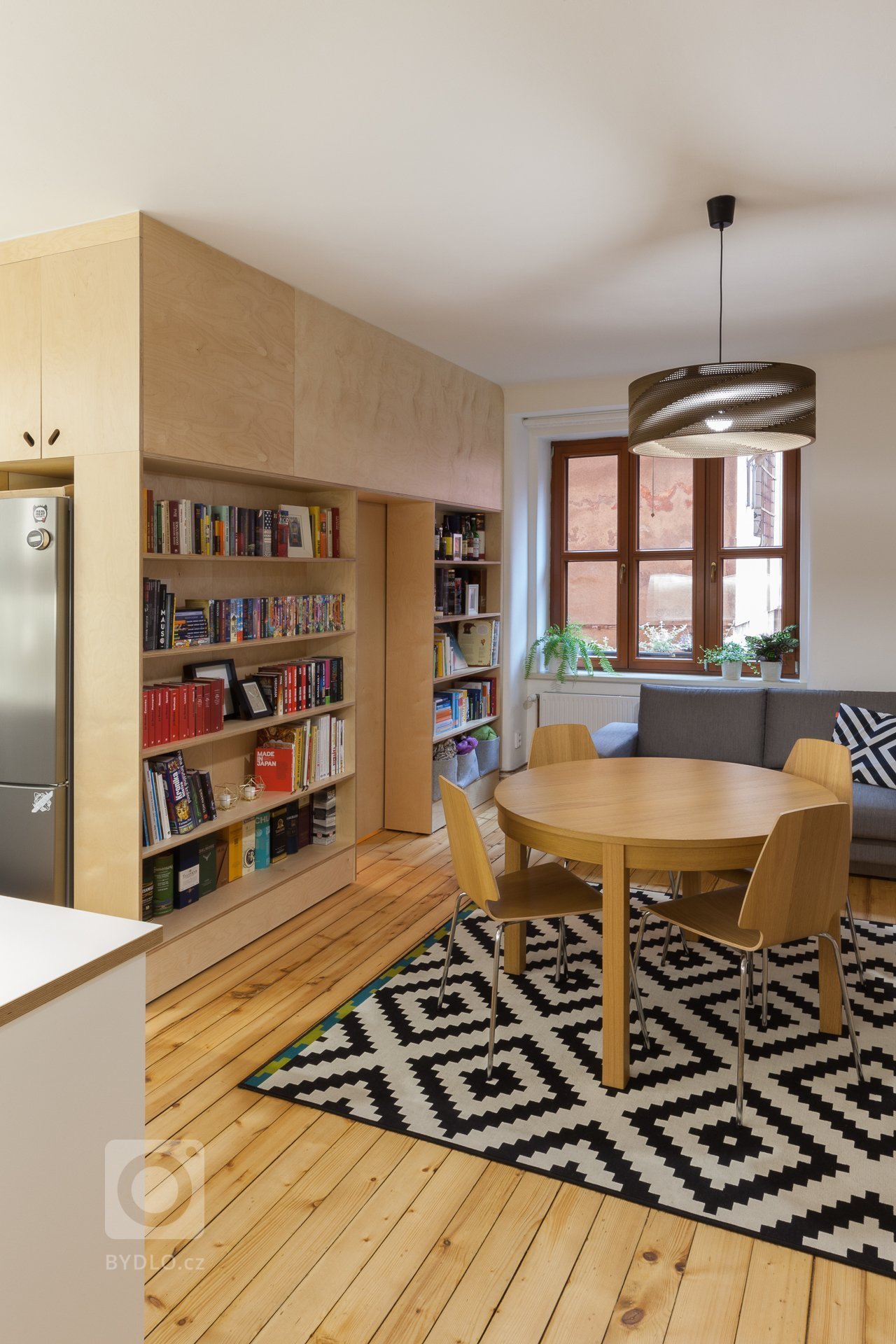 Nejdůležitější prostor, kde rodina tráví nejvíce času je obývací pokoj propojený s kuchyňským koutem a velkou knihovnou, která je integrovaná do vestavěné překližkové stěny, která odděluje nově vzniklý dětský pokoj. Jídelní stůl je umístěn centrálně. 