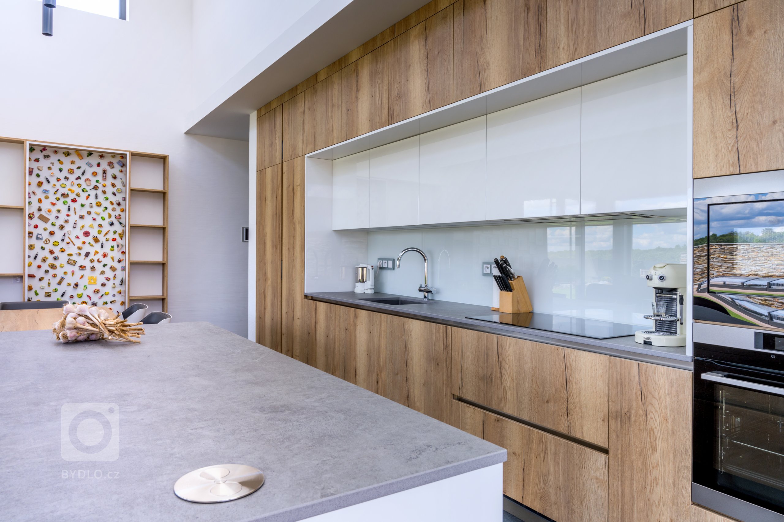 Velké dřevěné plochy sestavy působí čistě a romanticky, uprostřed jsou pak vystřídány mycím a varným centrem se skleněnou zástěnou, díky čemuž je prostor hezky symetrický i příjemně členitý.