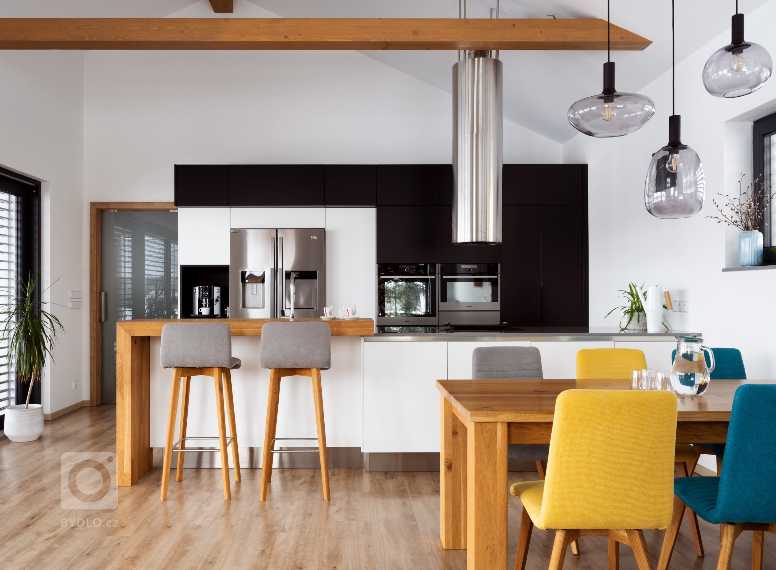 Hlavním stavebním prvkem této kuchyně je sestava vysokých skříní v kombinovaném provedení Evermatt bílý / Černý matt. Opticky se drží u zdi, uvnitř ovšem nabízí spoustu nespoutaného prostoru. Je osazena špičkovými spotřebiči - z nich do popředí vystupuje dvoukřídlá americká lednice. Před ní se pak nachází pracovní ostrůvek s pracovní deskou Technistone Noble Pro Cloud. Velkorysý solitér obsahuje jak mycí, tak varné centrum, na které od stropu dohlíží válcovitý odsavač, a je zakončen servírovacím pultíkem s posezením a vestavěnou vinotékou.