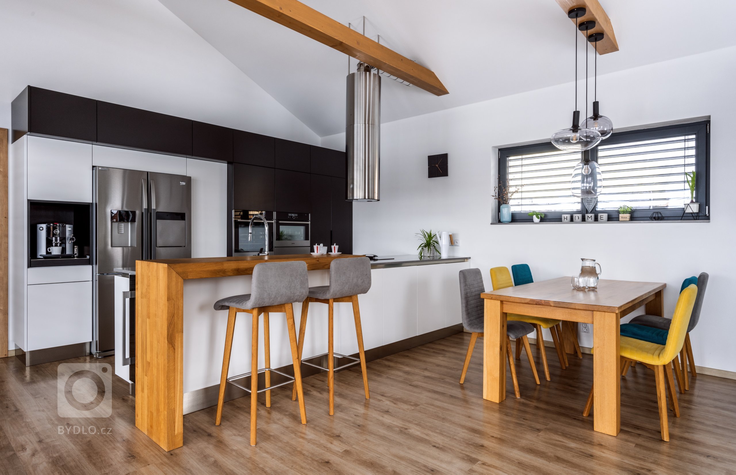 Hlavním stavebním prvkem této kuchyně je sestava vysokých skříní v kombinovaném provedení Evermatt bílý / Černý matt. Opticky se drží u zdi, uvnitř ovšem nabízí spoustu nespoutaného prostoru. Je osazena špičkovými spotřebiči - z nich do popředí vystupuje dvoukřídlá americká lednice. Před ní se pak nachází pracovní ostrůvek s pracovní deskou Technistone Noble Pro Cloud. Velkorysý solitér obsahuje jak mycí, tak varné centrum, na které od stropu dohlíží válcovitý odsavač, a je zakončen servírovacím pultíkem s posezením a vestavěnou vinotékou.