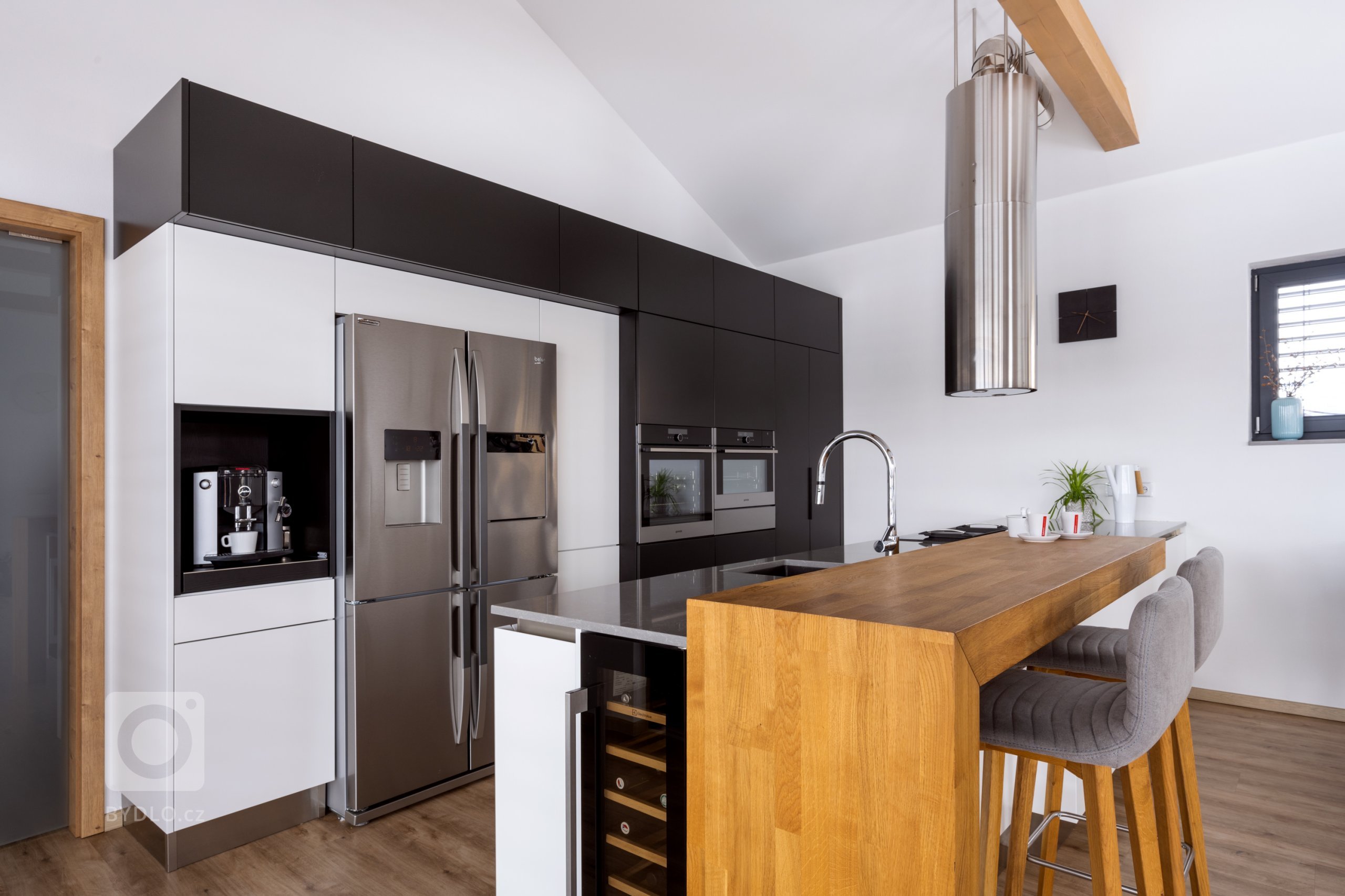 Hlavním stavebním prvkem této kuchyně je sestava vysokých skříní v kombinovaném provedení Evermatt bílý / Černý matt. Opticky se drží u zdi, uvnitř ovšem nabízí spoustu nespoutaného prostoru. Je osazena špičkovými spotřebiči - z nich do popředí vystupuje dvoukřídlá americká lednice. Před ní se pak nachází pracovní ostrůvek s pracovní deskou Technistone Noble Pro Cloud. Velkorysý solitér obsahuje jak mycí, tak varné centrum, na které od stropu dohlíží válcovitý odsavač, a je zakončen servírovacím pultíkem s posezením a vestavěnou vinotékou.