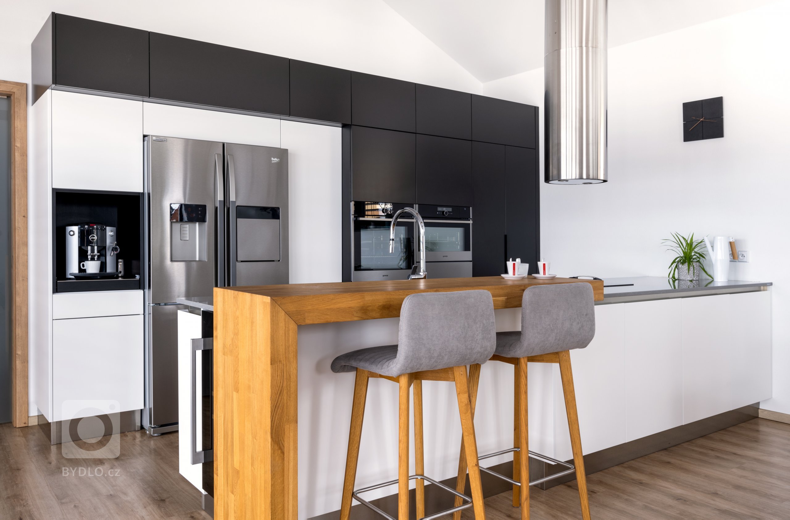 Hlavním stavebním prvkem této kuchyně je sestava vysokých skříní v kombinovaném provedení Evermatt bílý / Černý matt. Opticky se drží u zdi, uvnitř ovšem nabízí spoustu nespoutaného prostoru. Je osazena špičkovými spotřebiči - z nich do popředí vystupuje dvoukřídlá americká lednice. Před ní se pak nachází pracovní ostrůvek s pracovní deskou Technistone Noble Pro Cloud. Velkorysý solitér obsahuje jak mycí, tak varné centrum, na které od stropu dohlíží válcovitý odsavač, a je zakončen servírovacím pultíkem s posezením a vestavěnou vinotékou.