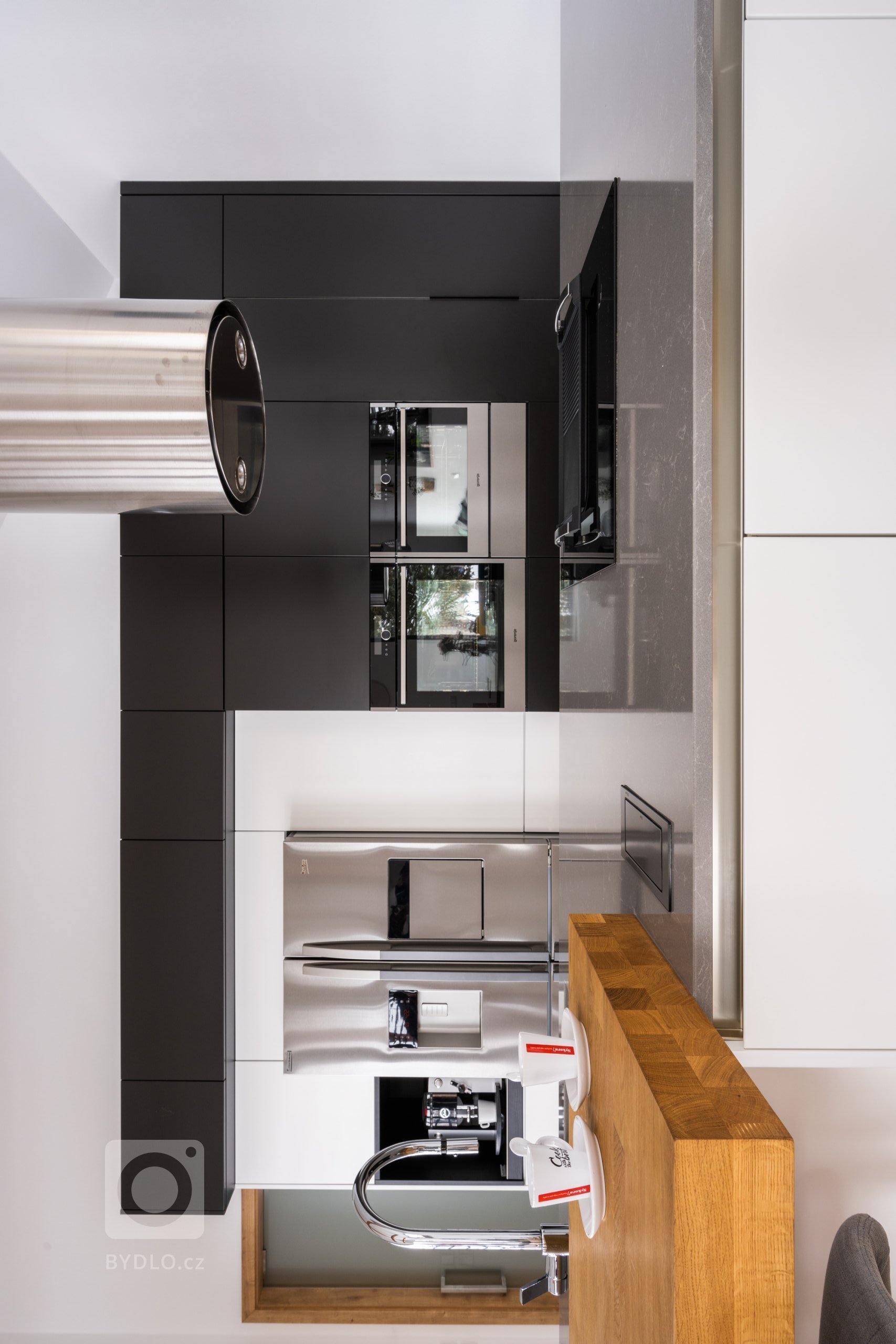 Hlavním stavebním prvkem této kuchyně je sestava vysokých skříní v kombinovaném provedení Evermatt bílý / Černý matt. Opticky se drží u zdi, uvnitř ovšem nabízí spoustu nespoutaného prostoru. Je osazena špičkovými spotřebiči - z nich do popředí vystupuje dvoukřídlá americká lednice. Před ní se pak nachází pracovní ostrůvek s pracovní deskou Technistone Noble Pro Cloud. Velkorysý solitér obsahuje jak mycí, tak varné centrum, na které od stropu dohlíží válcovitý odsavač, a je zakončen servírovacím pultíkem s posezením a vestavěnou vinotékou.
