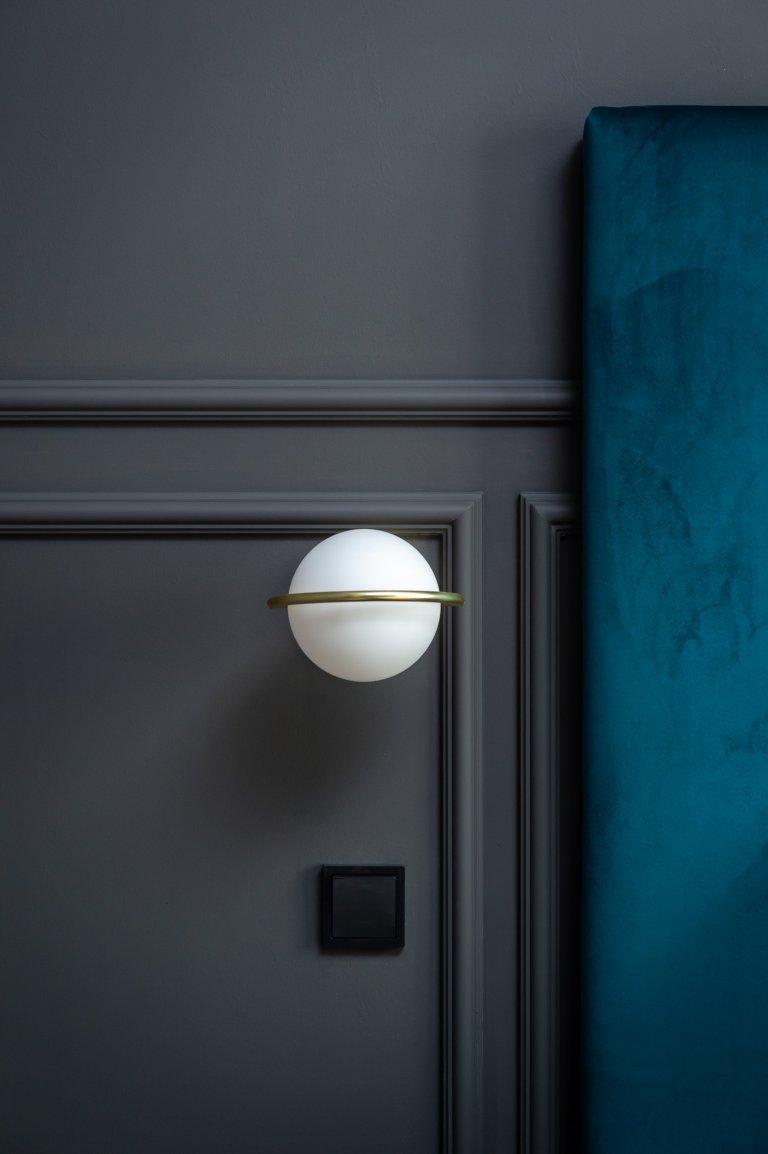 Detail v ložnici na opálové svítidlo v mosazném kroužku v pozadí antracitové stěny s okrasnými lištami.