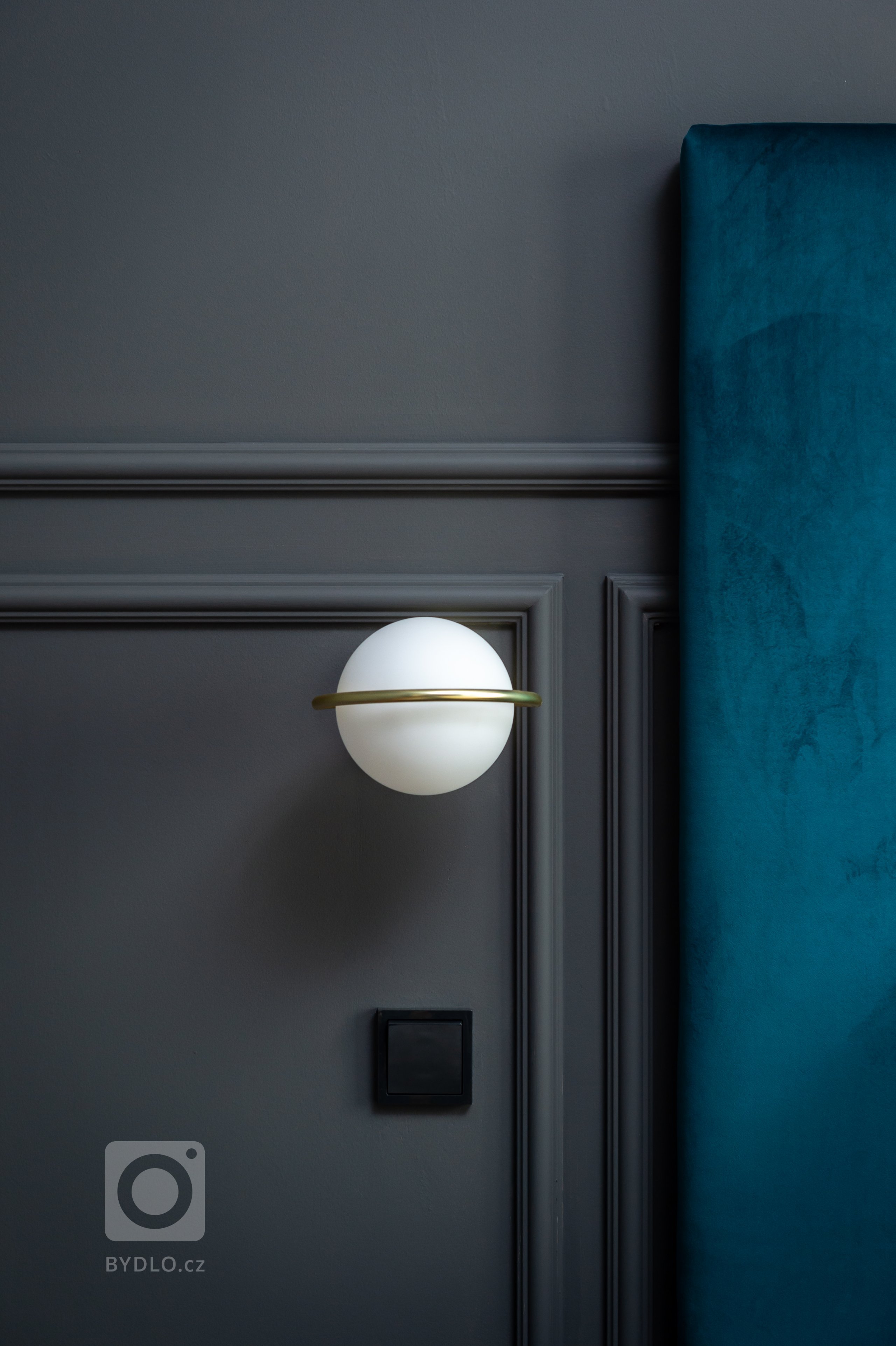 Detail v ložnici na opálové svítidlo v mosazném kroužku v pozadí antracitové stěny s okrasnými lištami.